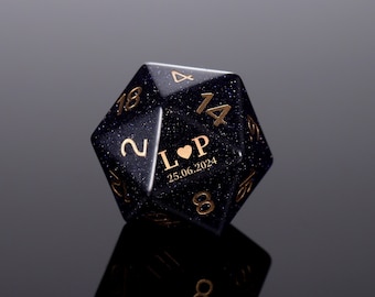 May include: A black, sparkly, 14-sided dice with gold numbers and a heart with the initials "L" and "P" and the date "25.06.2024".