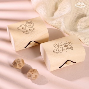 Può includere: Due scatole di legno color chiaro con coperchi curvi, con testo e disegni incisi. Una scatola dice "Date Night" e la data. L'altra scatola dice "Rebecca & James". Sono presenti anche due dadi di legno con vari simboli. Un tessuto color chiaro è sullo sfondo.