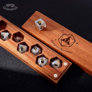 May include: Wooden dice box with a set of polyhedral dice. The dice are clear with black cat graphics and gold numbering. The box is engraved with the words "Dungeon Meowster" and a cat-themed geometric design.