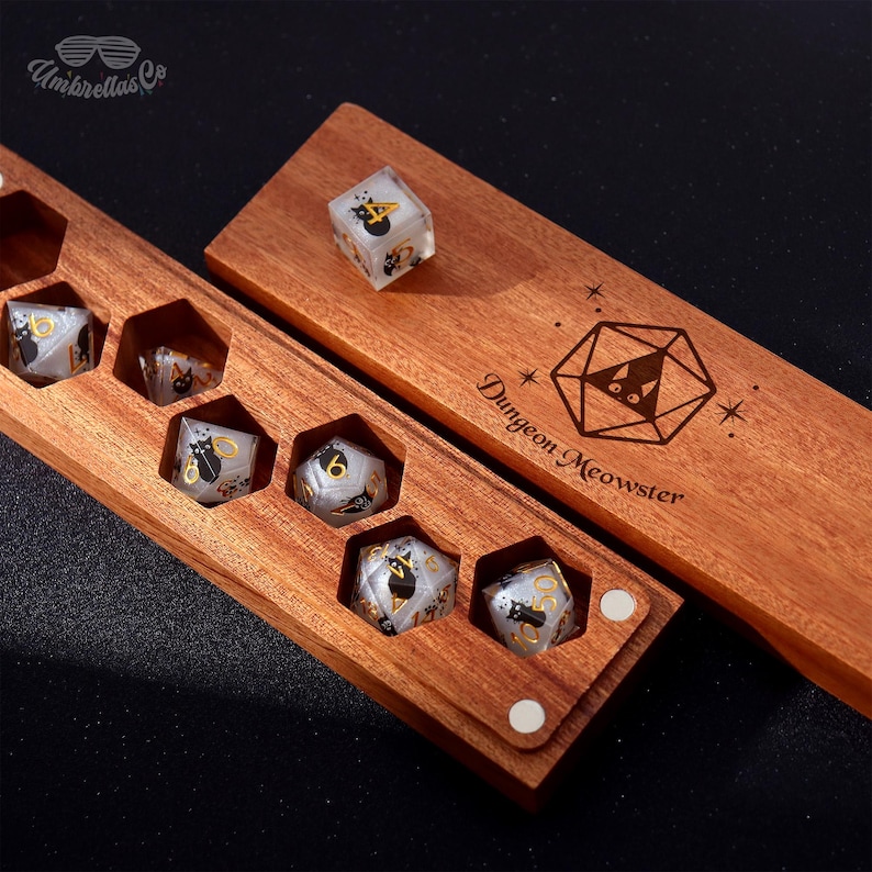 May include: Wooden dice box with a set of polyhedral dice. The dice are clear with black cat graphics and gold numbering. The box is engraved with the words "Dungeon Meowster" and a cat-themed geometric design.