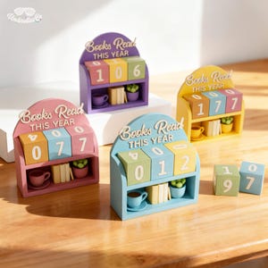 May include: A collection of colorful wooden book counters. Each counter displays the text "Books Read This Year" and features a shelf with a miniature cup, books, and a small plant. The counters are in various colors, including purple, pink, yellow, and blue.