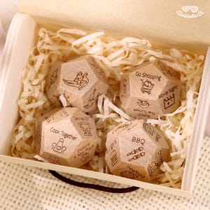 May include: A wooden box filled with four dodecahedron dice. Each die has engraved illustrations and text, including "Cook Together," "Go Shopping," and "Movie Night." The dice are nestled in shredded paper.