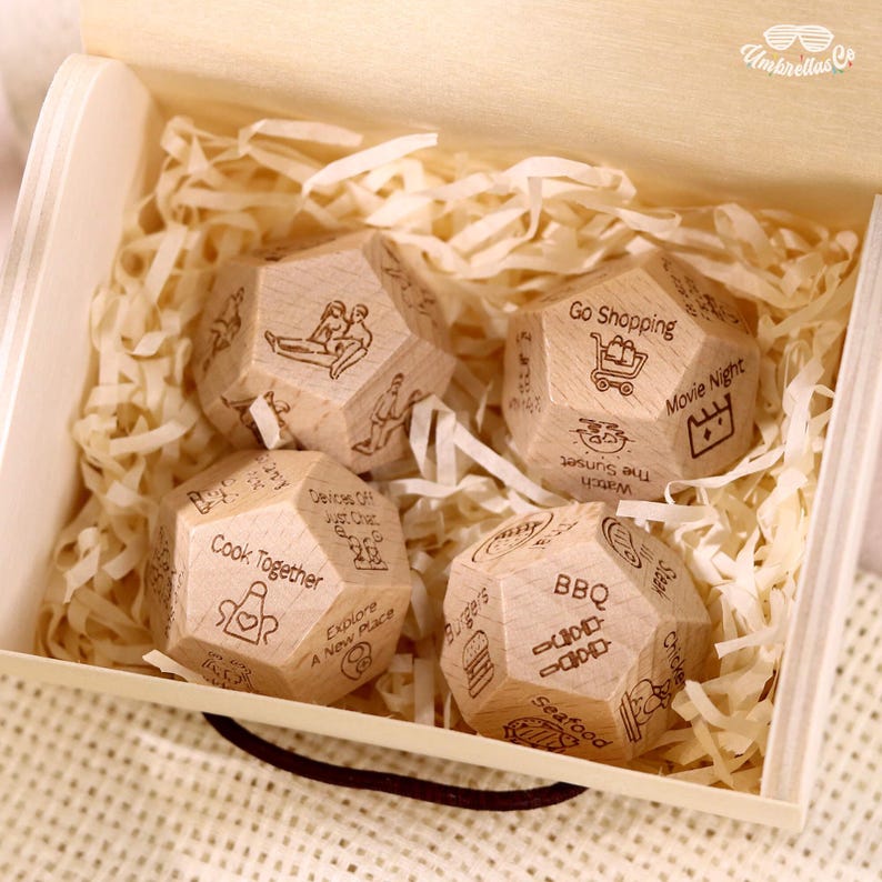 May include: A wooden box filled with four dodecahedron dice. Each die has engraved illustrations and text, including "Cook Together," "Go Shopping," and "Movie Night." The dice are nestled in shredded paper.