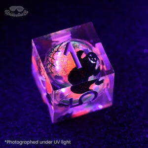 Könnte beinhalten: Ein klarer, würfelförmiger Kunstharzwürfel mit einem schwarzen Geist und der Zahl 1. Der Würfel hat ein leuchtendes orangefarbenes und grünes Innere, und der Text "Umbrellas Co" ist sichtbar. Das Bild ist mit "Photographed under UV light" beschriftet.