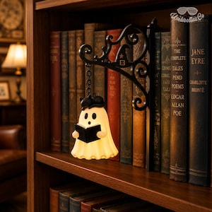 May include: A cream-colored ghost-shaped lamp with a black cat on its head, holding an open black book. The lamp hangs from a black metal wall bracket featuring scrollwork and a small witch hat icon, positioned between several vintage books.