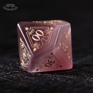 May include: A translucent, pinkish-purple, ten-sided die with gold-colored numerals and decorative designs. The die features the text "Umbrellas Co" in the upper left corner. The numbers 00, 10, 20, 30, 40, 50, 60, 70, 80, and 90 are displayed.