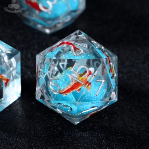 May include: Clear, multi-sided dice with silver numbering and a koi fish design. The dice feature a blue, glittery interior with a red and orange fish. The dice are for tabletop games.
