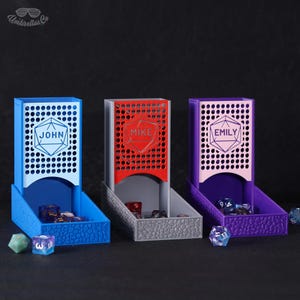 Op de afbeelding: Drie gepersonaliseerde dobbelstenentorens in blauw, rood en paars. Elke toren heeft een naam in een geometrische vorm op de voorkant. De torens hebben een geperforeerde bovenkant en een getextureerde basis, met dobbelstenen zichtbaar binnenin en eromheen.