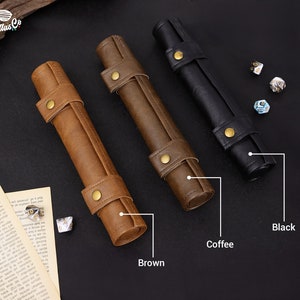 May include: Three leather dice rolling trays in brown, coffee, and black. The trays are rolled up and have a gold metal clasp. The trays are on a black surface with dice scattered around them.