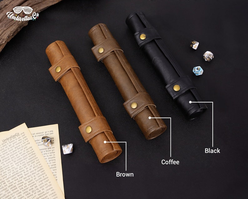 May include: Three leather dice rolling trays in brown, coffee, and black. The trays are rolled up and have a gold metal clasp. The trays are on a black surface with dice scattered around them.