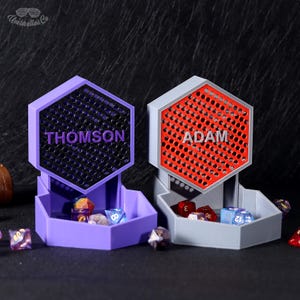 May include: Two purple and grey hexagonal dice towers with the names "THOMSON" and "ADAM" printed on the front. The towers have a honeycomb pattern and are filled with dice.