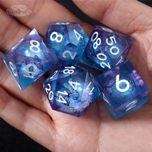May include: Set of six iridescent blue and purple dice with white numbers and butterfly designs. The dice are translucent and have a glittery finish.