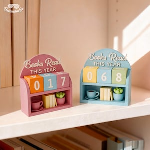 Op de afbeelding: Twee houten boekenleestellers. Eén is roze met oranje, blauwe en witte blokken met de tekst "017". De andere is blauw met gele, groene en witte blokken met de tekst "068". Elk heeft een plank met miniatuurboeken, een kopje en een plant.