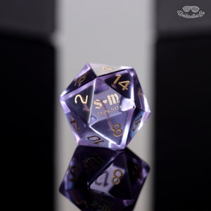 May include: A purple, clear, iridescent, 20-sided dice with gold numbers and the inscription "S+M 25.06.2024".