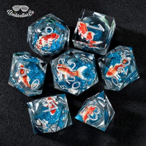May include: A set of seven polyhedral dice made of clear resin, each containing a miniature orange and white koi fish figurine suspended in blue glitter. The dice feature silver-colored numbers engraved on the surfaces, with the Umbrellas Co logo visible in the top left corner.