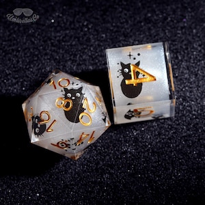 May include: Two dice, a twenty-sided die and a six-sided die, with a cat theme. The dice are clear with a glittery interior and gold numbering. Each die features a black cat illustration. The text "Umbrellas Co" is in the upper left corner.