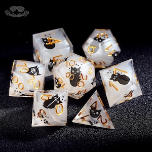 May include: A set of seven clear, glittery dice with black cat illustrations and gold numbering. The dice are various shapes, including a cube, a d20, and a pyramid. The dice are for tabletop games.
