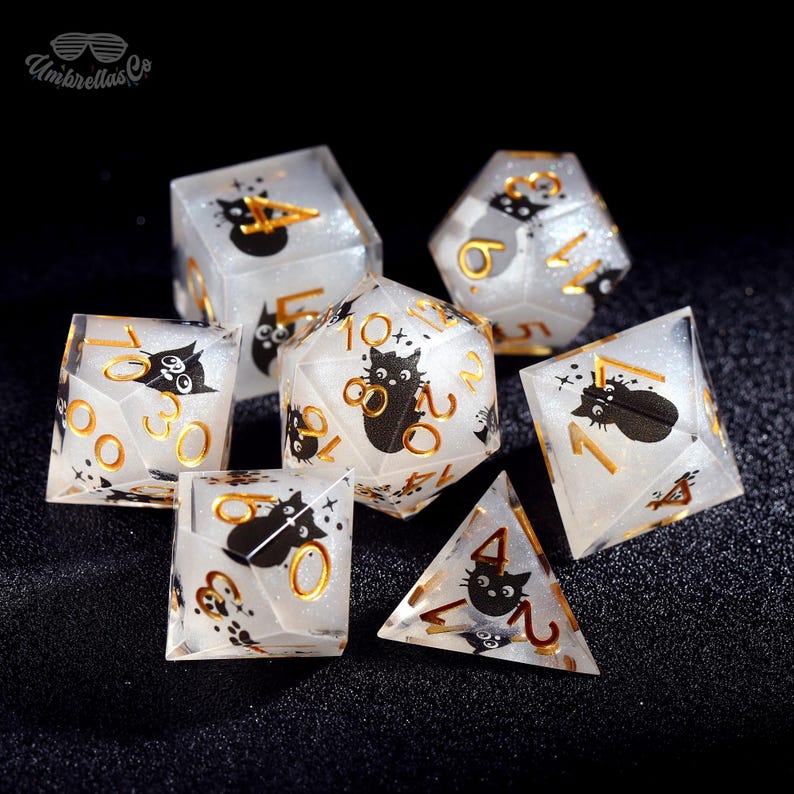 May include: A set of seven clear, glittery dice with black cat illustrations and gold numbering. The dice are various shapes, including a cube, a d20, and a pyramid. The dice are for tabletop games.