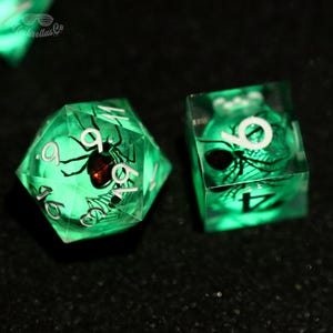 Puede incluir: Dos dados verdes brillantes con un diseño de araña y telaraña. Uno es un dado de 20 caras, el otro es un dado de seis caras. Los números son blancos y la araña es negra con un abdomen rojo. El texto "Umbrellas Co" está en la esquina superior izquierda.