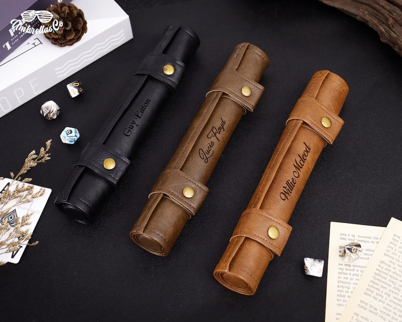 May include: Three personalized leather dice rolling trays. The trays are rolled up and have a brown leather exterior with gold accents. The trays are labeled with the names "Guy Eaton", "Lucie Floyd", and "Willie McLeod".