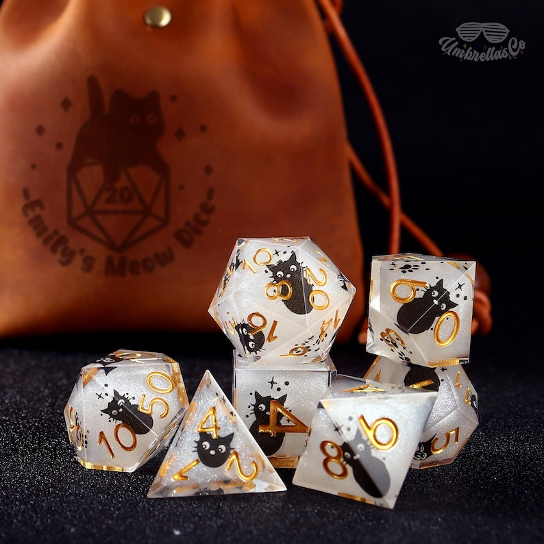 May include: A set of polyhedral dice with a cat theme. The dice are clear with gold numbering and black cat illustrations. A brown leather bag with a cat logo and text is in the background.