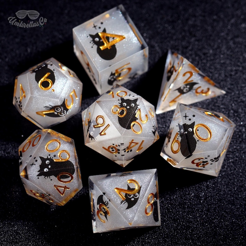 May include: A set of seven clear, glittery dice with black cat illustrations and gold numbering. The dice are various polyhedral shapes, including a d20, d12, d10, d8, d6, and d4. The text "Umbrellas Co" is in the upper left corner.