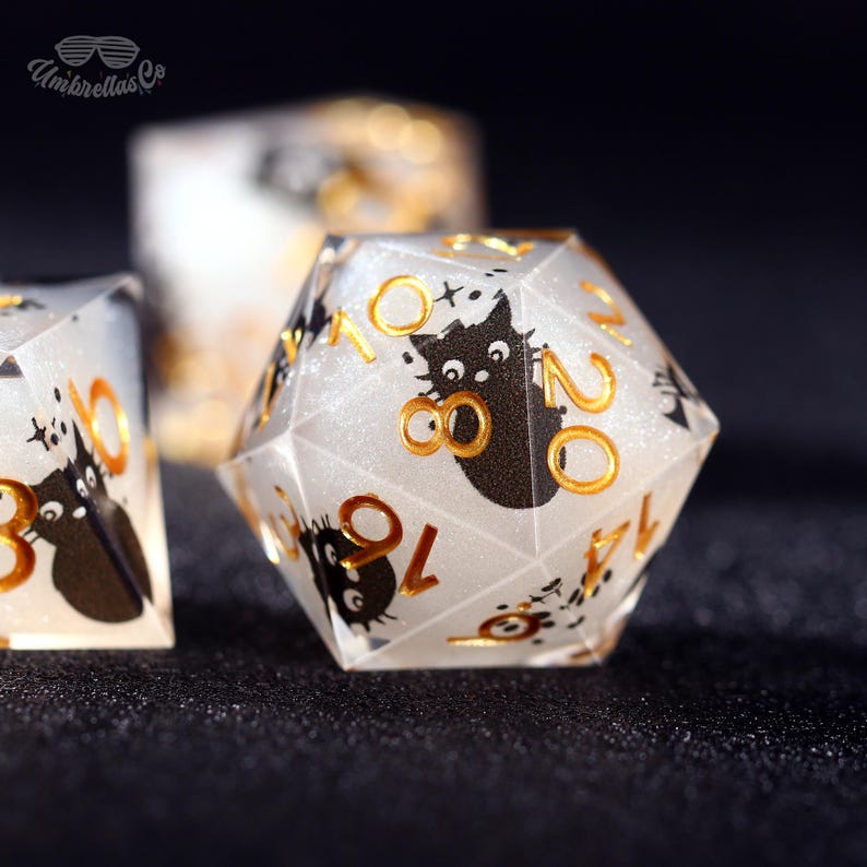 May include: Close-up of a set of polyhedral dice. The dice are clear with a glittery interior and feature black cat illustrations and gold numbering. The dice are for tabletop games.