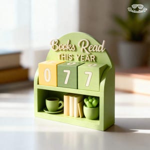 May include: A green wooden calendar in the shape of a bookshelf, with the words "Books Read THIS YEAR" on top. The calendar shows the number "077" on three rotating blocks. The shelf holds a small teacup, books, and a succulent.
