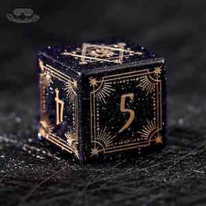 May include: A dark blue, glittery dice with gold-colored markings and numbers. The dice features intricate sunburst designs around the numbers and symbols. The text "Umbrellas Co" is visible in the upper left corner.