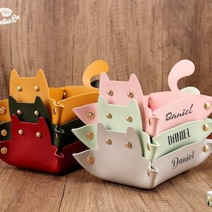 May include: Set of five colorful cat-shaped leather trays, stacked on top of each other. The top tray is light gray and has the name "Daniel" written on it. The trays are perfect for organizing small items.