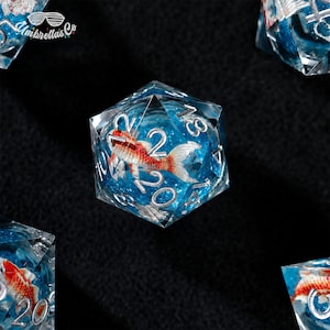 Op de afbeelding: Een twintigzijdige veelvlakdobbelsteen van heldere hars met een miniatuur oranje en witte koikarperfiguur die in blauwe glittervloeistof zweeft. De dobbelsteen heeft zilverkleurige gegraveerde cijfers en het Umbrellas Co logo.