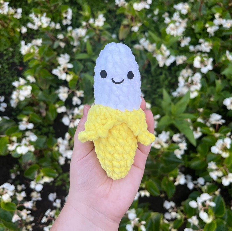 May include: A crocheted banana plush toy with a white face and a happy expression. The banana is yellow and has a white stem.