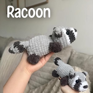 May include: Two crocheted raccoon plush toys. The raccoons are gray with dark gray and white accents on the face and tail. The text "Raccoon" is displayed in white above the toys.