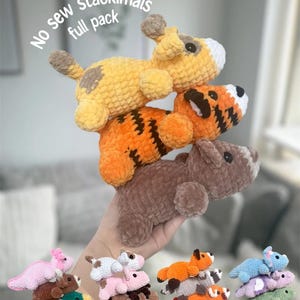 Op de afbeelding: Een stapel handgemaakte gehaakte dierenspeeltjes in geel, oranje en bruin. Het bovenste speeltje is een gele giraf met bruine vlekken, gevolgd door een oranje tijger en een bruine beer. De tekst "No sew stackimals full pack" is zichtbaar.