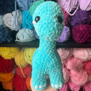 Puede incluir: Un juguete de dinosaurio de crochet azul turquesa con ojos negros y pies blancos. El juguete est&aacute; siendo sostenido por una mano frente a un estante de hilo de colores.