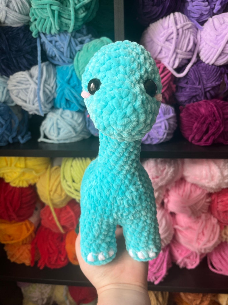Puede incluir: Un juguete de dinosaurio de crochet azul turquesa con ojos negros y pies blancos. El juguete est&aacute; siendo sostenido por una mano frente a un estante de hilo de colores.