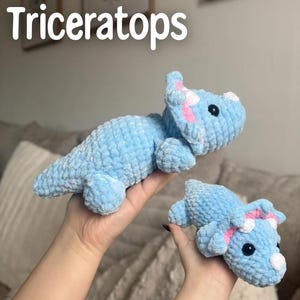 May include: Two crocheted triceratops dinosaurs in light blue with pink and white accents. The larger dinosaur is lying down, while the smaller one is held in a hand. The word "Triceratops" is written in white above the dinosaurs.