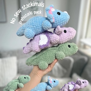 May include: A stack of three crocheted dinosaur plush toys in blue, purple, and green, with a smaller green, blue, and purple dinosaur plush in the foreground. The text "No sew stackimals prehistoric pack" is in the background.