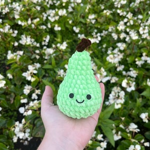 May include: A green crocheted pear with a brown stem and a smiling face. The pear is being held in a hand against a background of white flowers.