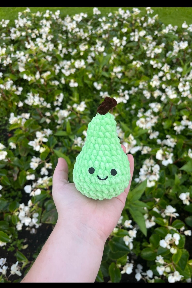 May include: A green crocheted pear with a brown stem and a smiling face. The pear is being held in a hand against a background of white flowers.