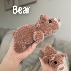 May include: Two plush, brown teddy bears. The larger bear is lying down, and the smaller bear is held in a hand. The bears have black eyes and white accents on their snouts. The word "Bear" is written in white text.
