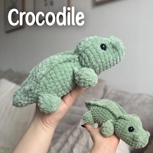 May include: Two crocheted green crocodile plush toys. The larger crocodile is lying down, and the smaller one is upright. Both have black button eyes and are made of a soft, textured yarn. The word "Crocodile" is written in white above the toys.