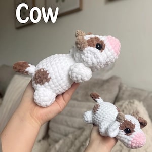 May include: Two crocheted cow plushies. The larger cow is white with brown spots, ears, and tail, and a pink nose. The smaller cow has the same design. The word "Cow" is written in white above the larger cow.
