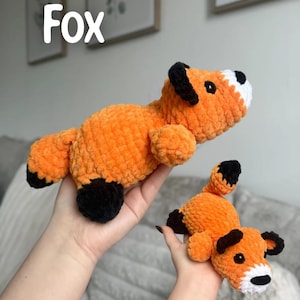 May include: Two orange crocheted fox stuffed animals. The foxes have black ears, paws, and tail tips, with white accents on the face. The word "Fox" is written in white block letters at the top of the image.