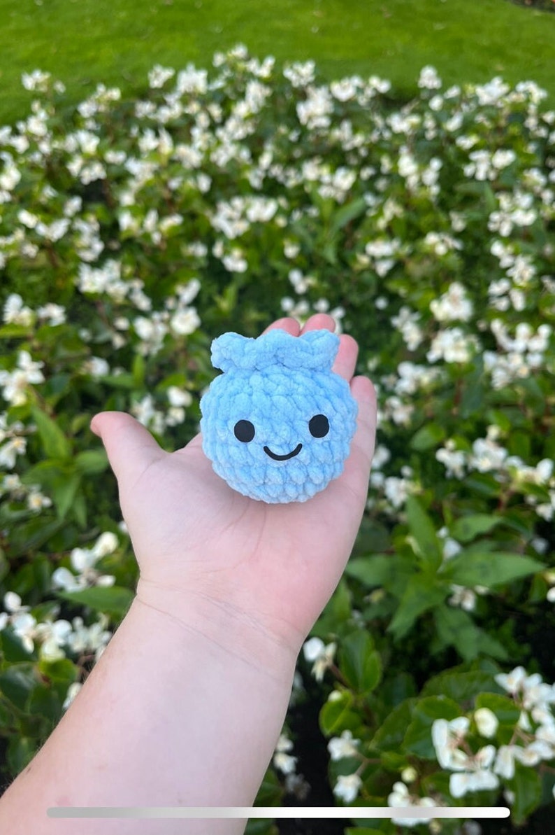May include: A small, light blue crocheted plush toy shaped like a blueberry with a smiling face. The toy is being held in a person's hand.