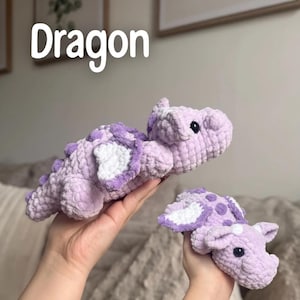 May include: Two crocheted dragon plushies in shades of lavender and white. The larger dragon has a long body with a spiky back and the smaller one has wings. The word "Dragon" is written in white across the top of the image.