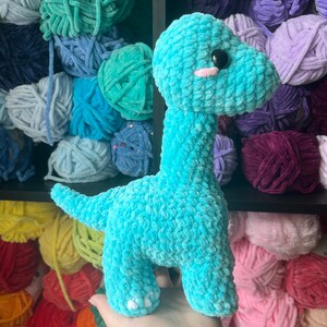 Puede incluir: Un juguete de dinosaurio de crochet azul turquesa con pies blancos. El juguete est&aacute; sentado en un estante con ovillos de lana de colores.