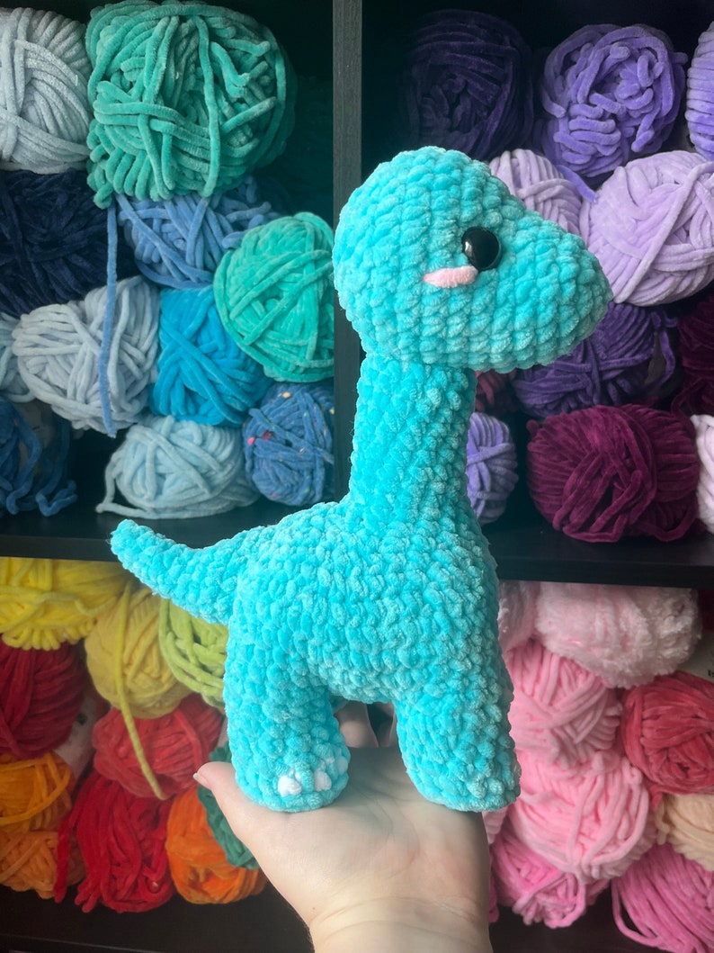 Puede incluir: Un juguete de dinosaurio de crochet azul turquesa con pies blancos. El juguete est&aacute; sentado en un estante con ovillos de lana de colores.