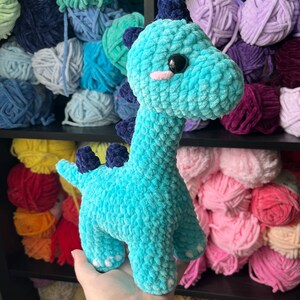 Puede incluir: Un juguete de dinosaurio de crochet azul turquesa con ojos blancos y una nariz rosa. El juguete est&aacute; siendo sostenido por una mano frente a un estante de hilo de colores.
