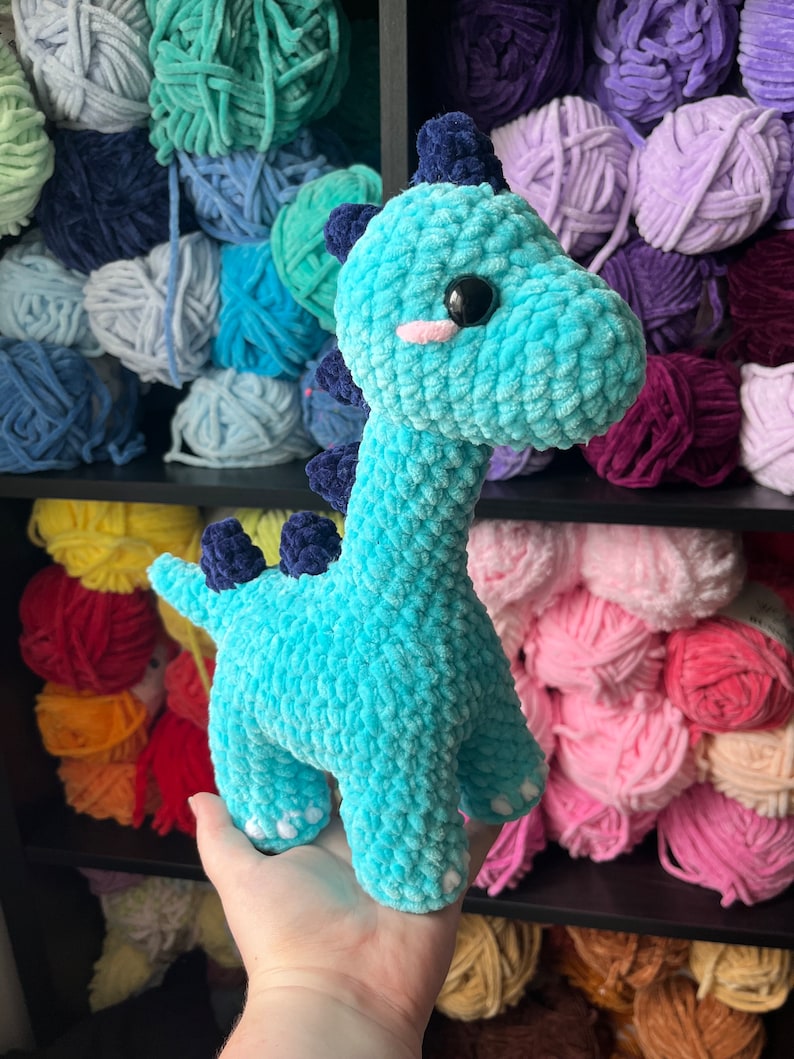 Puede incluir: Un juguete de dinosaurio de crochet azul turquesa con ojos blancos y una nariz rosa. El juguete est&aacute; siendo sostenido por una mano frente a un estante de hilo de colores.